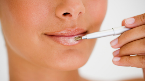 Metade do rosto de uma mulher, que está segurando um pincel de gloss e aplicando nos lábios.