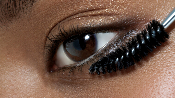 Imagem está focado apenas em um dos olhos de uma mulher que está segurando um aplicador de rímel próximo aos cílios.