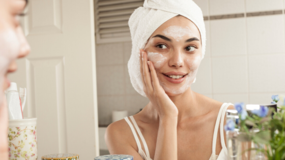 Uma mulher olhando para o espelho, usando uma toalha na cabeça e aplicando um produto facial de cor esbranquiçada.
